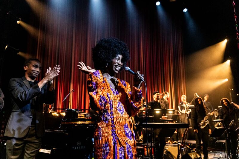 A singer is smiling and waving her hand as she holds a microphone, there's a band in the background and a man behind her applauding