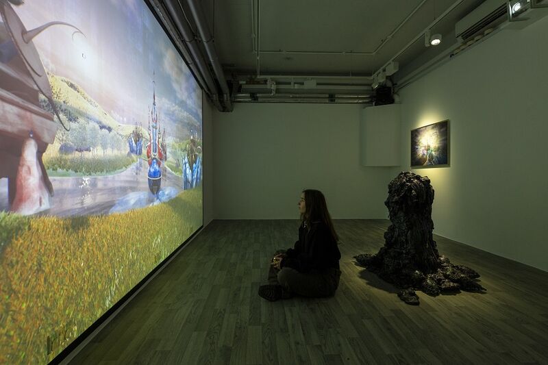A girl sits on the floor, behind her is an oozing sculpture, and in front of her is a big screen