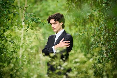 A white man in a suit stands amongst lots of green foliage