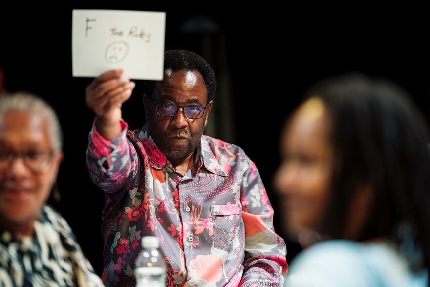 A black man in a colourful shirt holds up a piece of white paper with a sad face on it. It is a bit blurry but the letter F is visible. There are two other people sitting in front of him, both blurry, but they appear to be smiling.