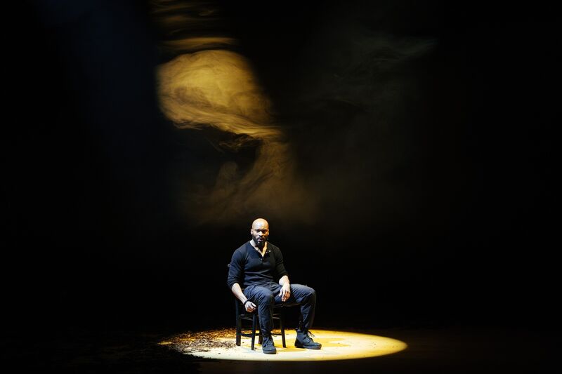 Arinze, a black man, is sat on a black chair lit with a yellow spotlight, there is a yellow circle on the floor and a yellow haze in the air above him