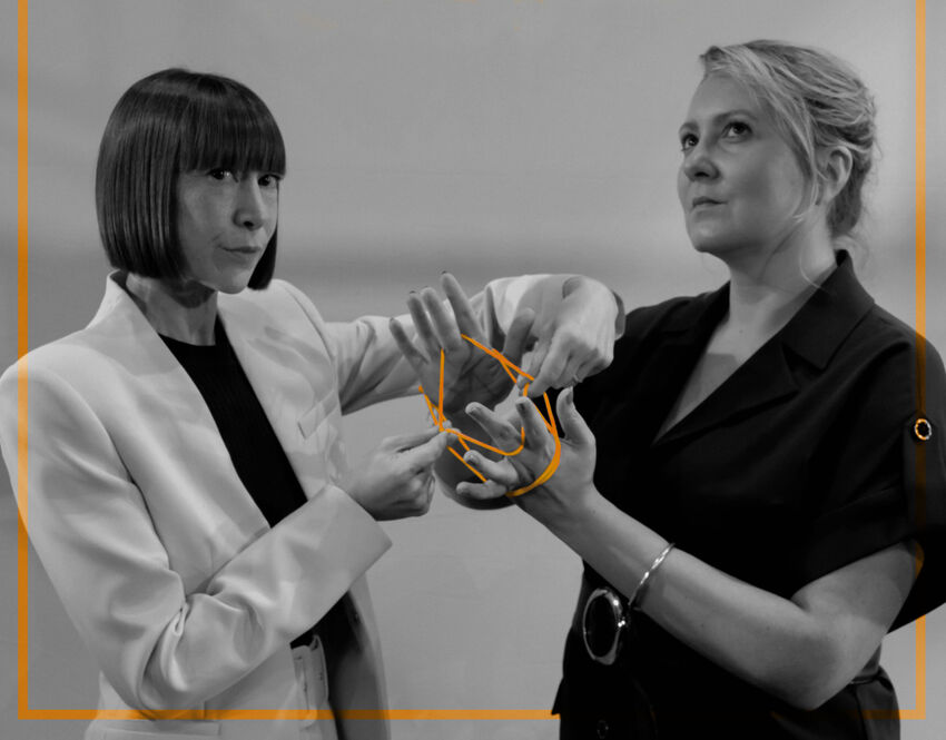Two people stand playing cats cradle with orange string