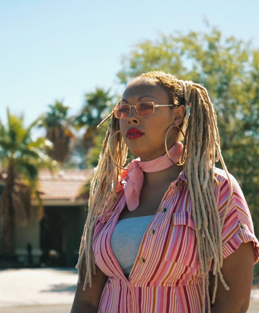 Harleighblu with blond dreads in pigtails with pink glasses, a pink scarf tied in her neck, large gold hoop earings and a pink stripy shirt. A blue sky and palm trees out of focus behind her