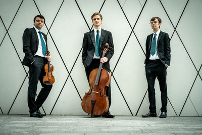 Mithras Trio standing in front of a white wall with black diamond outlines. One member of the trio is holding a violin, and another holding a cello and another with his hands in his pocket. They are all wearing grey suits and blue ties