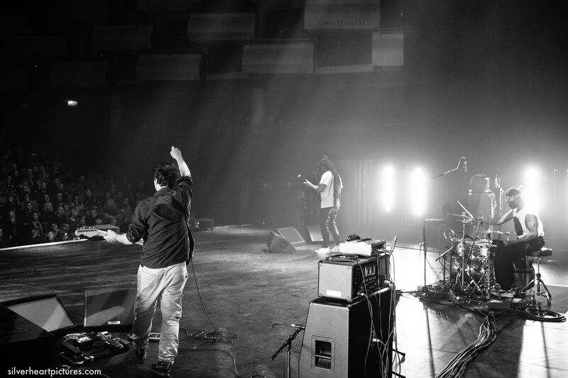 Black and white photo taken behind three musicans on stage. On the left is the bass player, central is the guitarist and on the right is the drummer