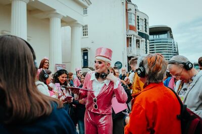 Alfie Ordinary in drag with a pink sequin cropped jacket, trousers and pilllbox hat, holding a clipboard and headphones on their neck, a crowd around them wearing headphones