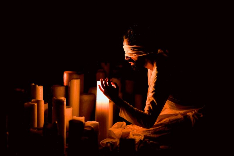 A man is kneeling, surrounded by candles, he holds one hand up toward the candles. He is wearing a white blindfold and a white top.