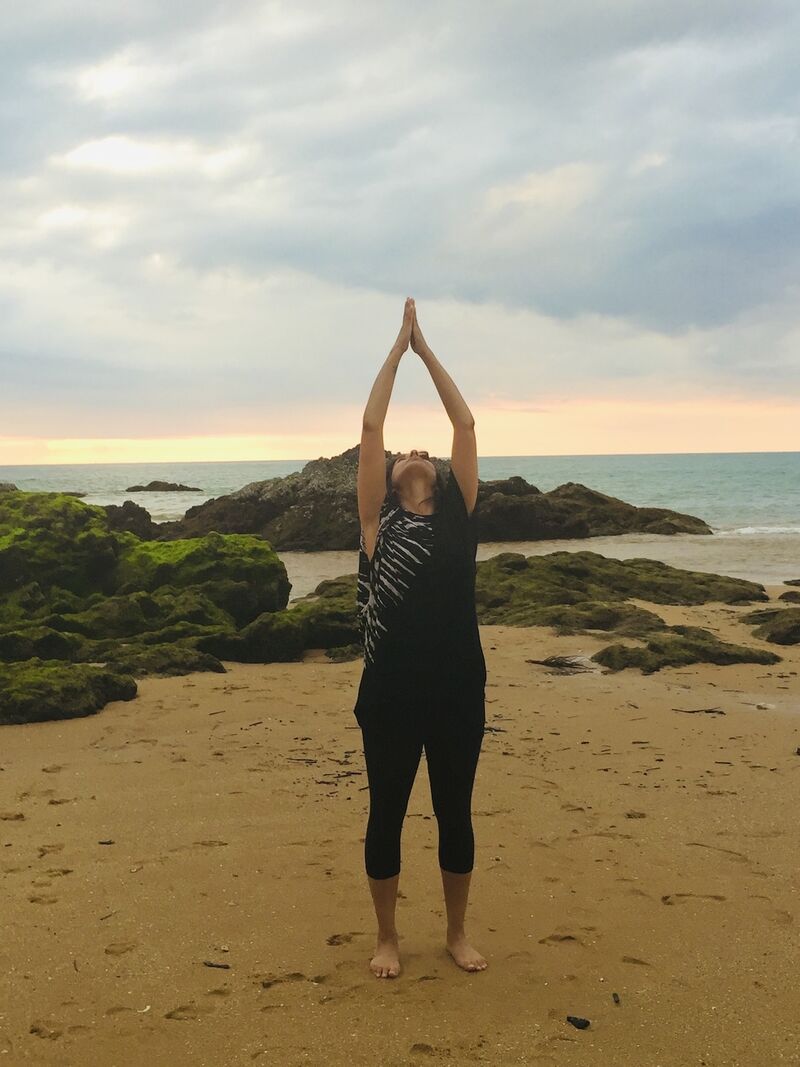 A woman stands on a beach. Her hands reach into the air with her palms together in a prayer position and she looks up towards her hands