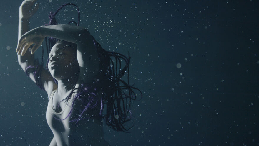 A dancer with long purple hair floats underwater with their arms above their head