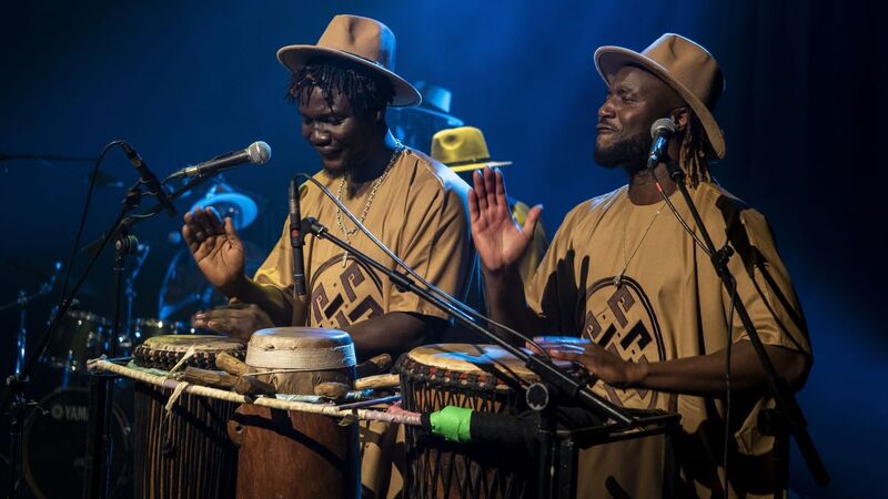 Two drummers play Djembe drums, wearing matching sand-colour t-shirts and hats