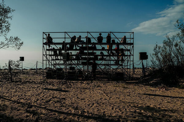 An audience stand is silhouetted against a late afternoon sky. It is placed on a beach area, and there are around twenty people spread along the different levels of the stand in a variety of poses. Several loudspeakers on stands are visible. The location is Bolderaja, in Riga, Latvia.