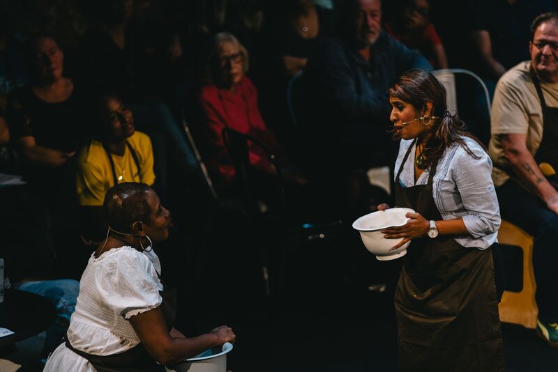 A woman sing to another woman sitting down. Both hold mixing bowls and wear cooking aprons