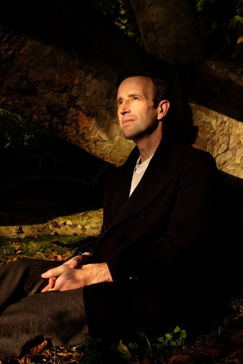 Robert Macfarlane, a white man, sits amongs some tree branches