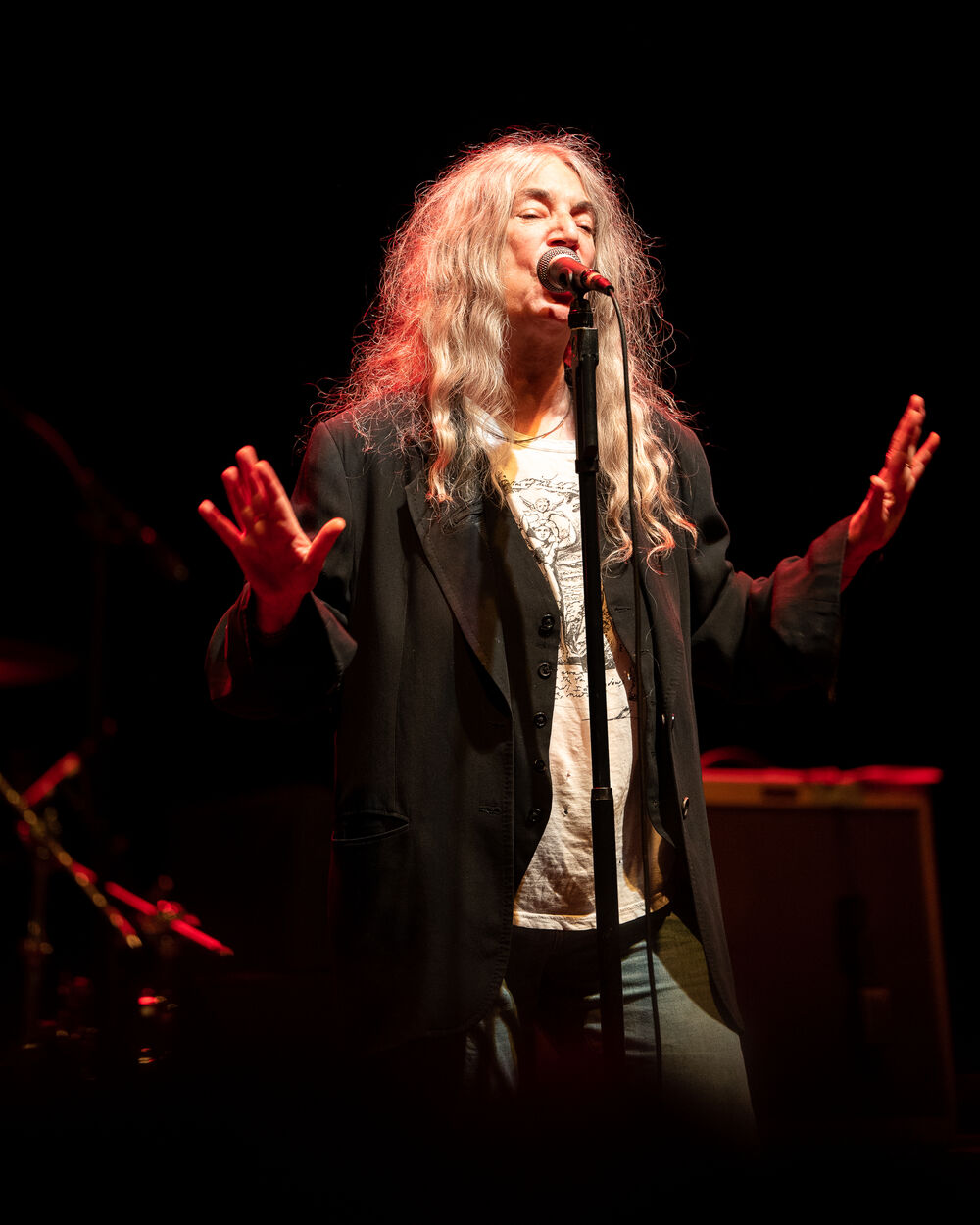 A lady in her 70s with long wavy grey hair wearing a black blazer sings into a microphone with both hands held up in front of her