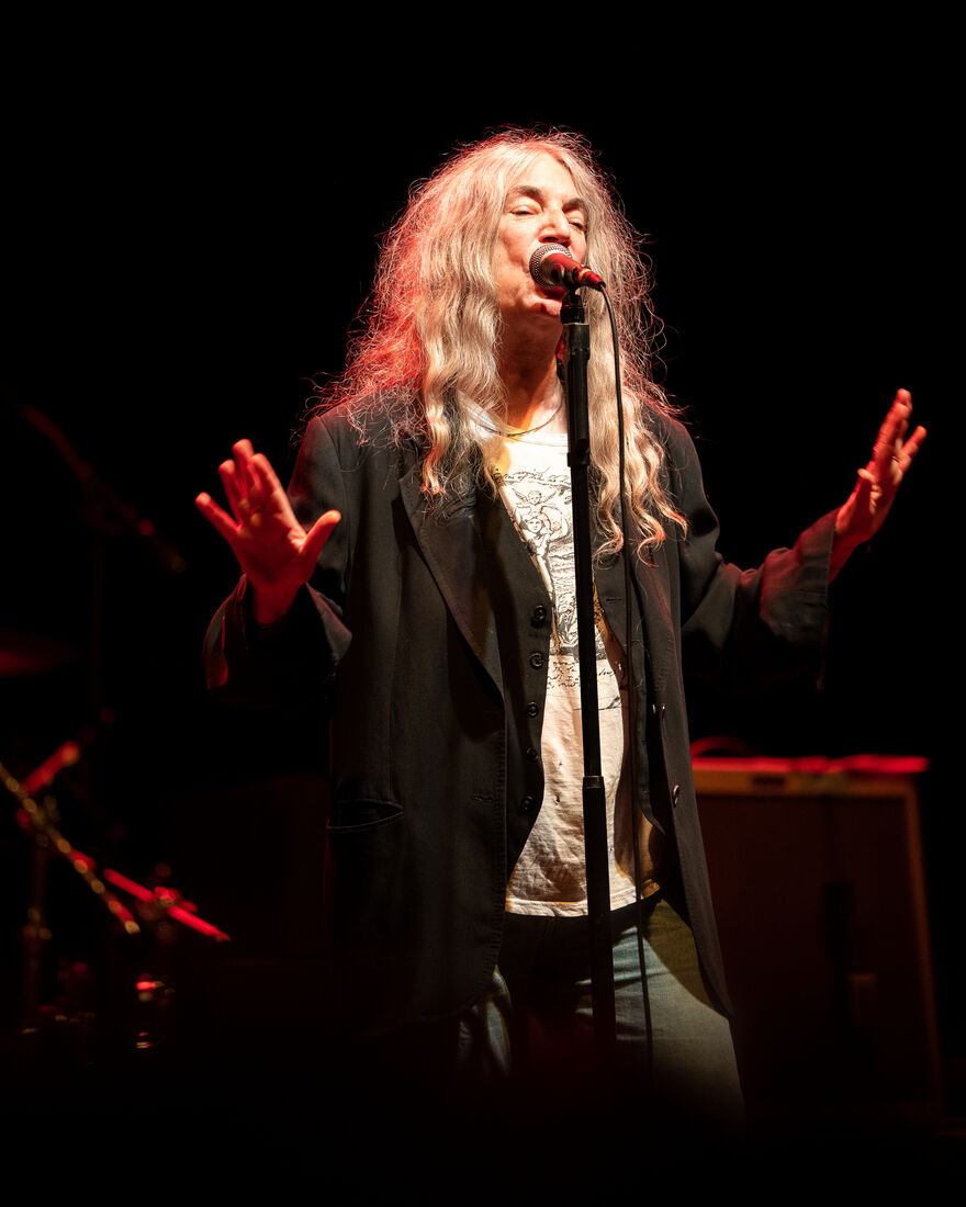 A lady in her 70s with long wavy grey hair wearing a black blazer sings into a microphone with both hands held up in front of her