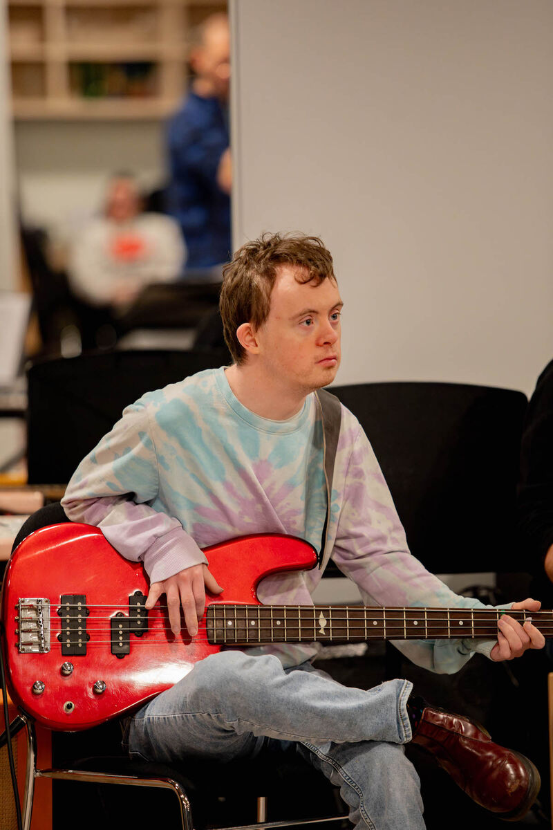 An Orchestra 360 musician sits and plays a bright red bass guitar. He is wearing a tie-dye jumper.