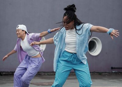 Two people, one dressed in light blue, the other in light purple, are dancing, their arms outstretched
