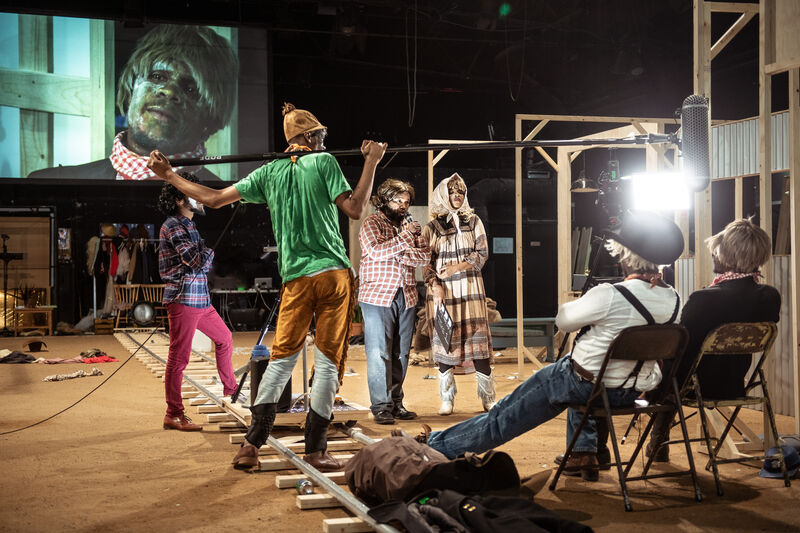 A boom mic is held above to people dressed as cowboys as they're sat down and facing away from the camera beside a wooden railway track