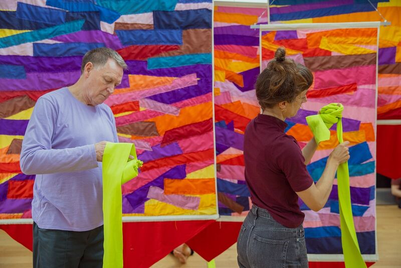 Standing against a brightly coloured tapestry, a man and a woman hold long yellow pieces of fabric, examining them closely