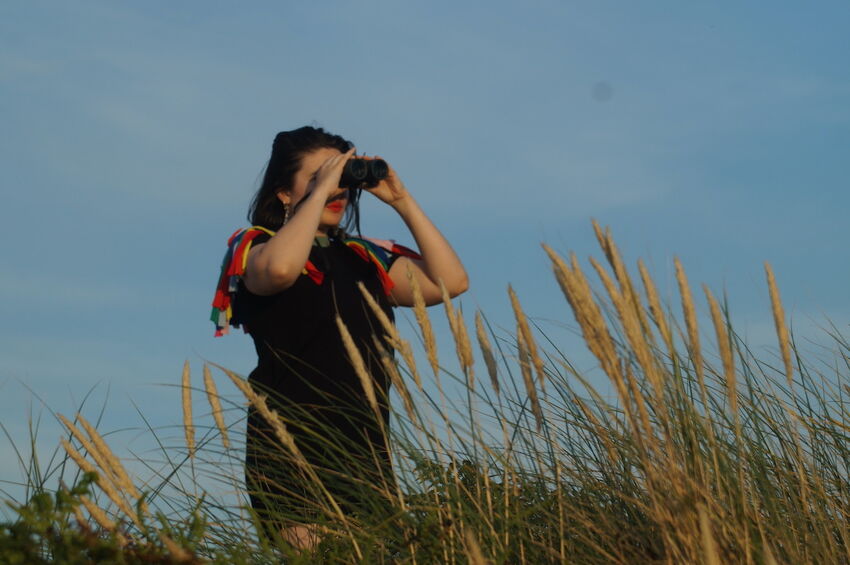 Tilly Ingram is dressed in a black top with colourful strips of fabric at her shoulders reminiscent of bird wings. She holds binoculars up to her eyes