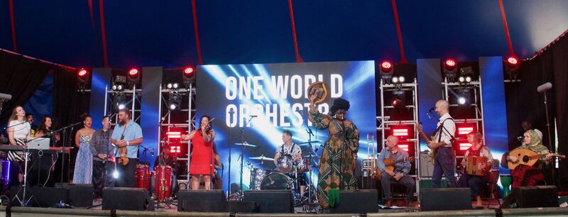 The musicians from One World orchestra perform on a stage. They're all holding instruments or singing into microphones
