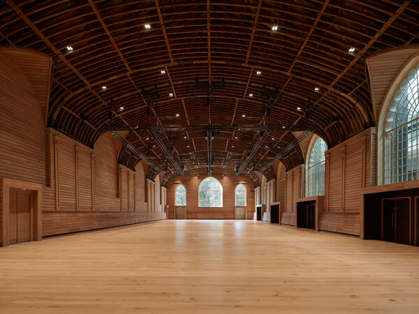 A large room with wooden floors and walls and an exposed beam ceiling, at the end and along the right hand side of the room are large arched windows