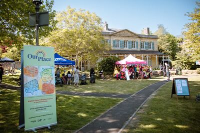 A large house and gardens with some stalls set up in front. In the foreground is a placard advertising Our Place