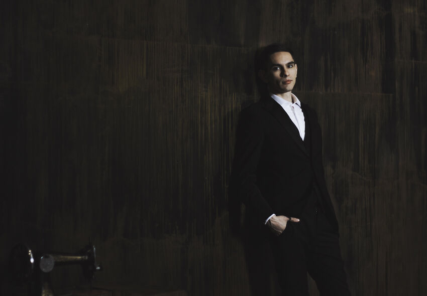 Dmitry Shishkin waring a black suit and white shirt, leaning against a dark wall