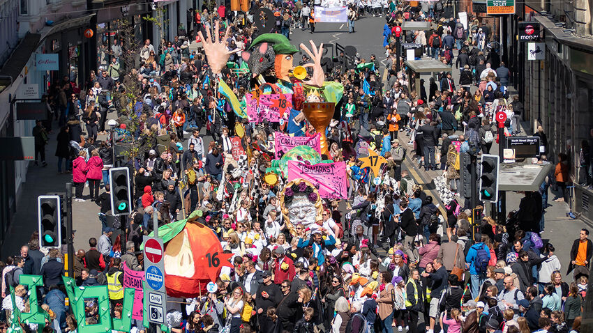 A bright, colourful parade makes its way along a road. The focal point is a large puppet which towers above the parade participants