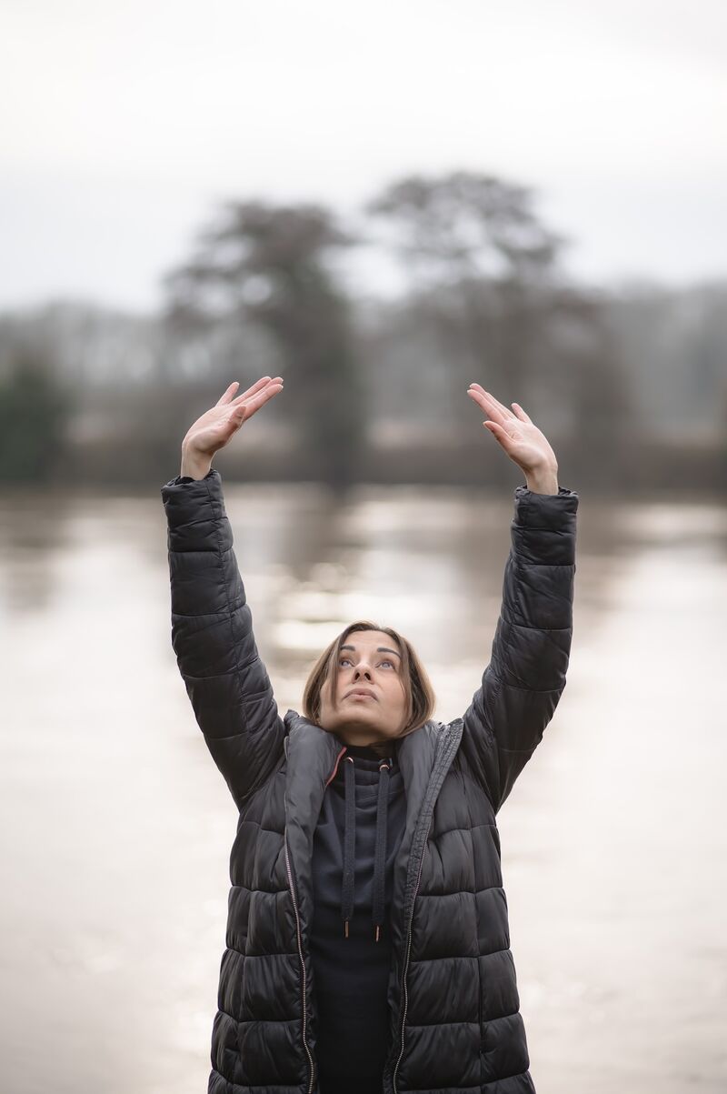 A woman is stood by a lake. She is dressed in black sports clothes and reaches her hands arms into the air. She gazes towards her outstretched hands with a calm expression