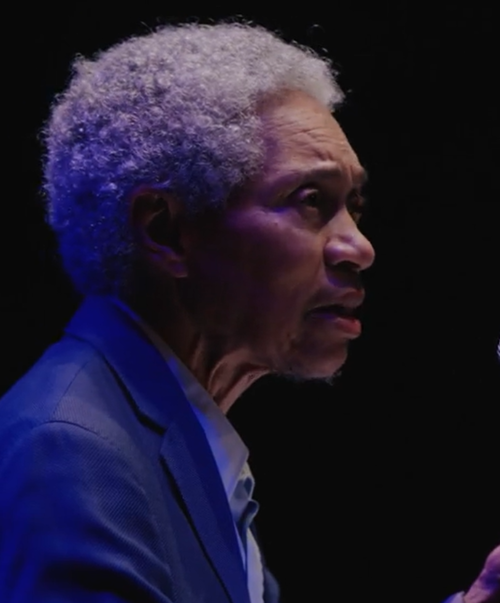 A side portrait of a black elderly man with a short grey afro wearing a suit and singing out towards an audience