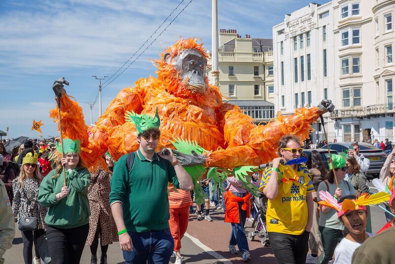 A group of people are parading along the sea front. They are holding a giant orangutan sculpture