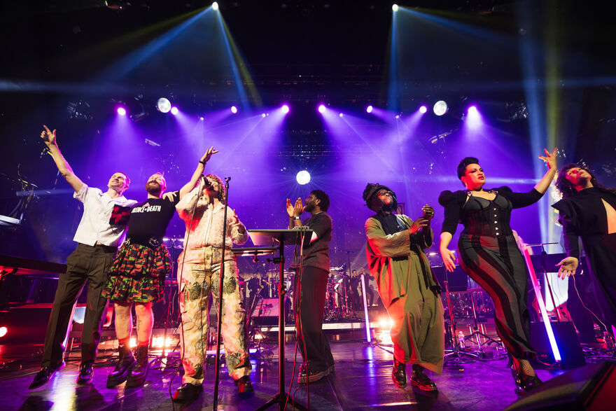 A group of performers stand on stage. They are happy and smiling, with a live band playing behind them.