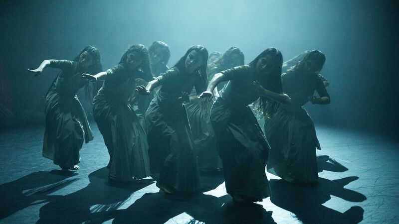 A group of female dancers stand in close formation under blue hued lighting, bending over with their arms reaching out