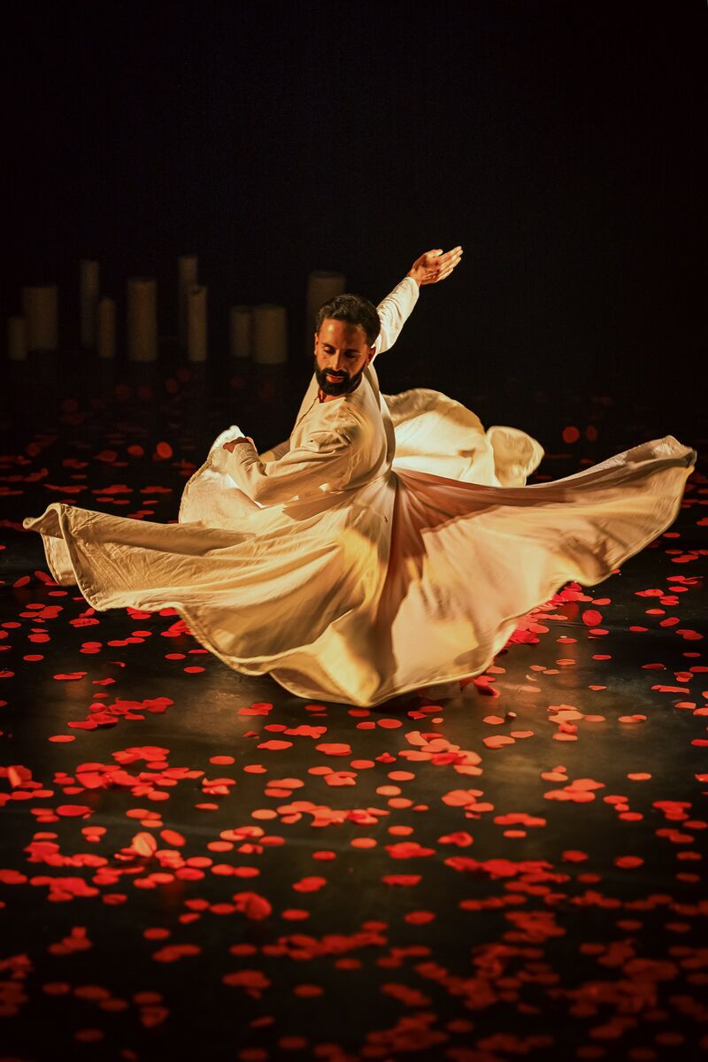 A man in a white dress, appears to be spinning low to the floor, which is covered in red petals