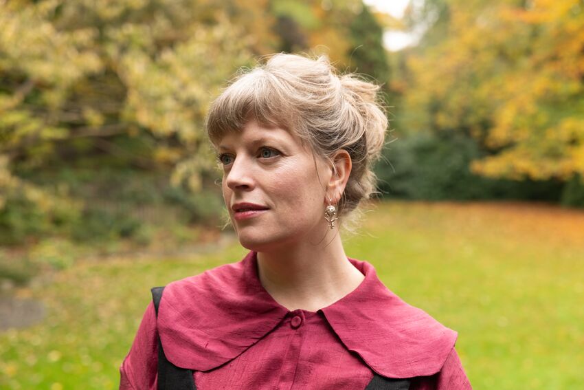 Sarah Perry is a white woman with blonde hair tied up in a bun, wearing a purple blouse and pearl earrings. She's standing outside in front of a background of trees