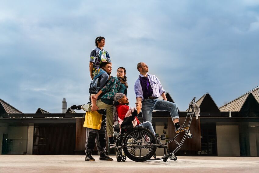 five dancers pose in front of a building with a clear blue sky. They are all gathered around a dancer who uses a wheelchair and wear brightly coloured costumes