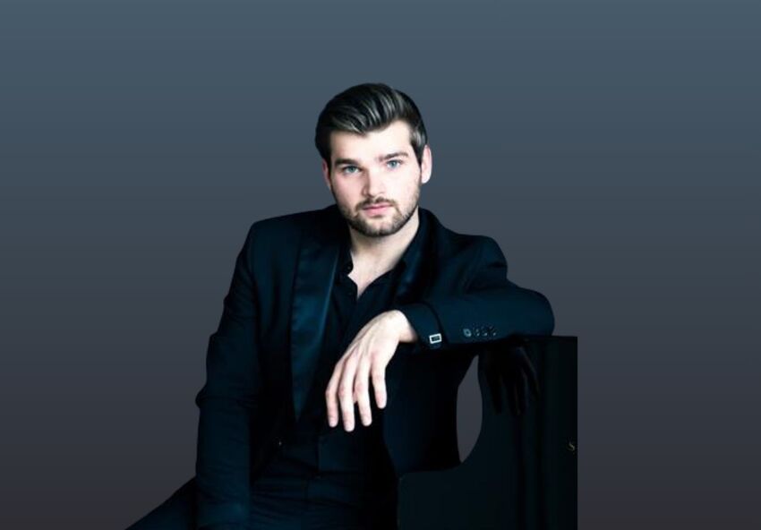 Rafael Kyrychenko wearing a navy blue suit leading on a piano. He is staring at the camera and sitting in front of a dark blue background