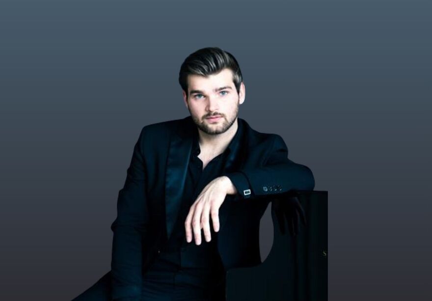 Rafael Kyrychenko wearing a navy blue suit leading on a piano. He is staring at the camera and sitting in front of a dark blue background