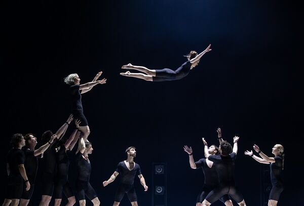 On the left, a group of acrobats are holding one of their members high up, she has flung another acrobat over to another group who are ready and wating to catch her
