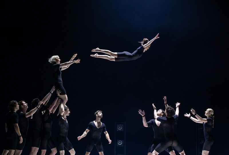 On the left, a group of acrobats are holding one of their members high up, she has flung another acrobat over to another group who are ready and wating to catch her