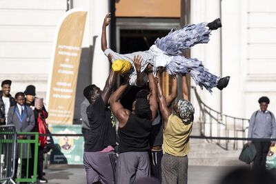 A man in feathered trousers and a yellow beanie hat is held aloft by another group of men