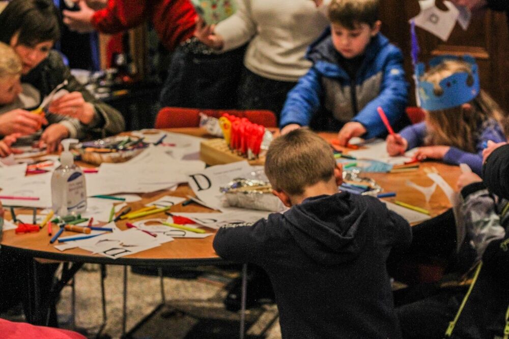 A group of young people sit around a craft table. They are busy making things and are surrounded by craft materials. One of them wears a paper crown that they have made