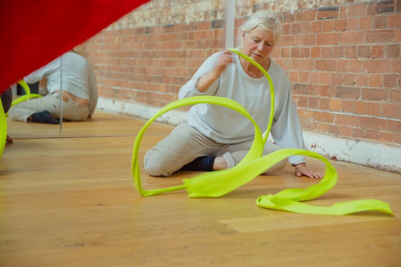 An older woman with grey hair sits on a wooden floor. She is wearing all white and spinning a long yellow piece of material