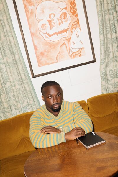 A portrait of Jason Okundaye. He wears a stripy green and orange jumper and sites on a mustard coloured sofa. A book is on the table in front of him