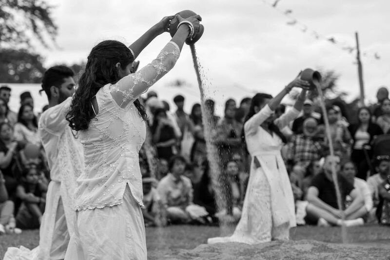 Three performers kneel on the ground in white lace, their hands above their heads tipping pots of sand on the ground