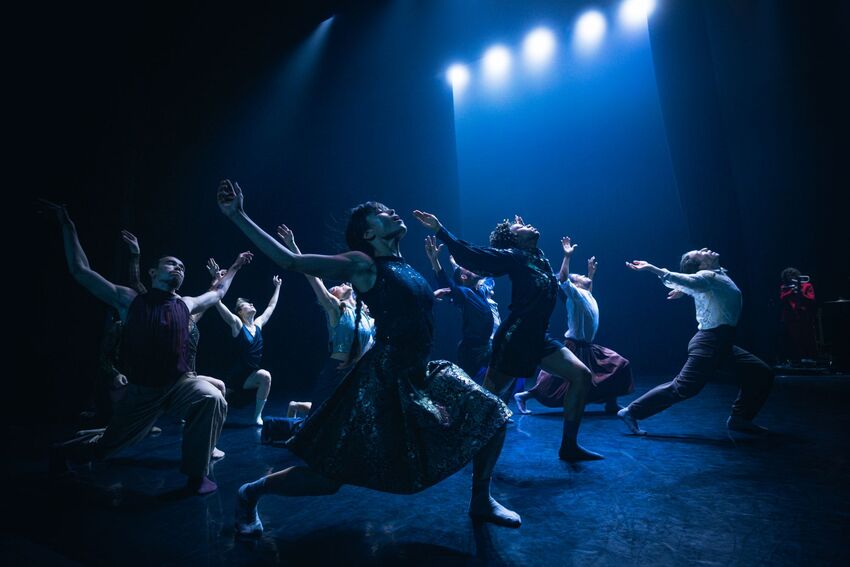 A group of dancers lunge forward and throw their arms backwards, intense blue lighting surrounds them