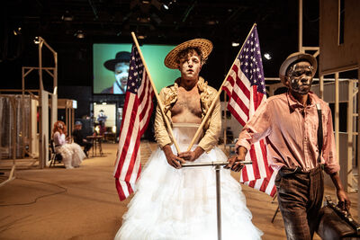 Two people in costumes with painted faces stand indoors; one wears a white tulle skirt with a cream jacket, hat, and holds two American flags, while the other wears a shirt, suspenders and hat.