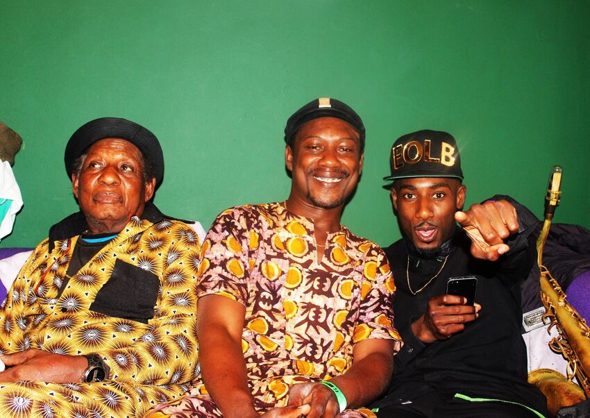 Three men sat on a sofa next to a saxophone. The man in the middle is smiling, on his right is an older man in a lively patterned suit, looking less happy. To the left is a younger man, wearing a cap and pointing forwards.
